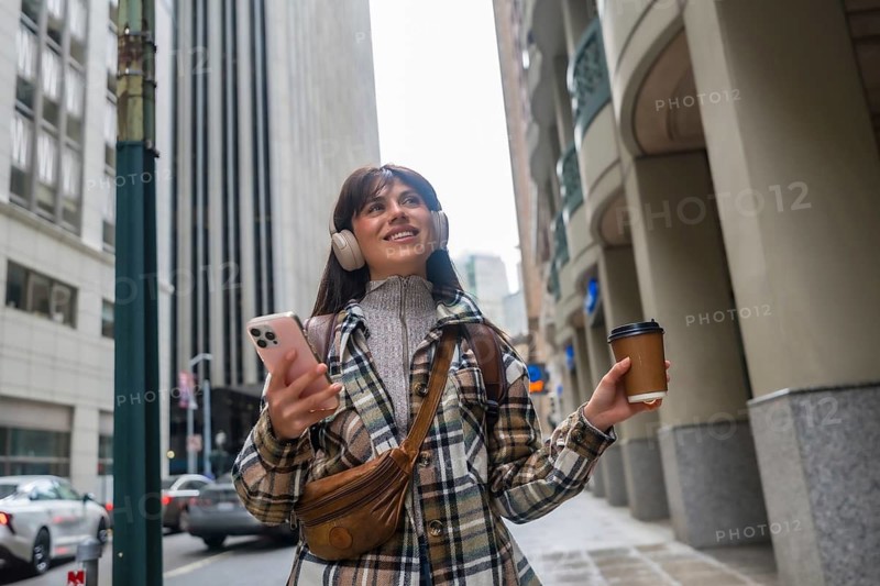 Young woman walking along a san francisco urban street, wearing headphones, smiling while looking up, holding a smartphone and a takeaway coffee cup, reflecting a modern city lifestyle