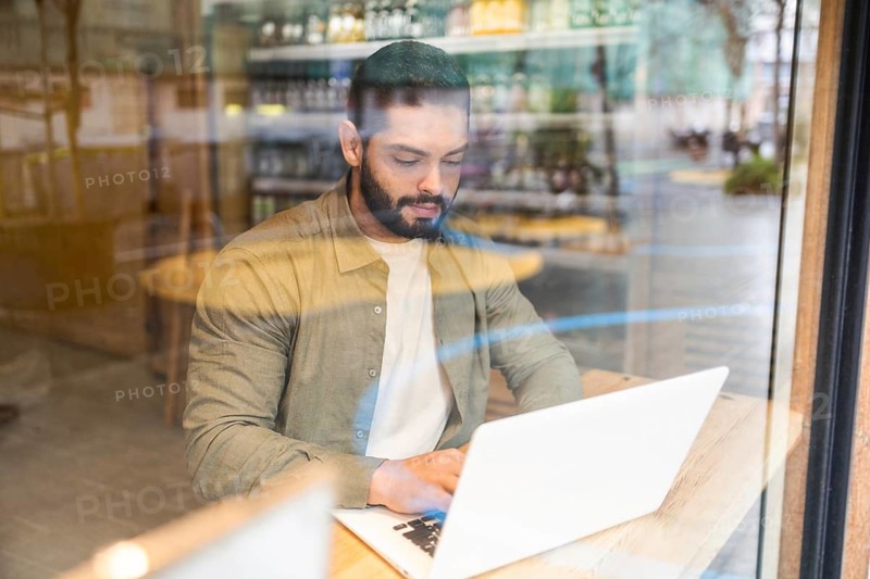 A man works on a laptop in a modern cafe, viewed through a glass window. The setting offers a glimpse into urban life, with reflections creating an interesting visual effect