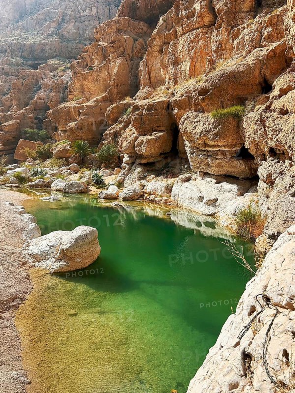 A tranquil oasis in Wadi Ash Shab, Oman, featuring emerald green waters surrounded by rugged rocky cliffs and sparse vegetation