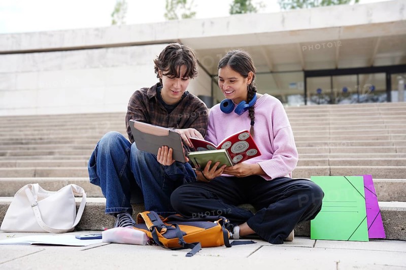 A non-binary teen and a female teen sit on school steps, engrossed in study materials. They are surrounded by backpacks and books and a tablet, symbolizing a return to school and collaboratively engaging in educational activities