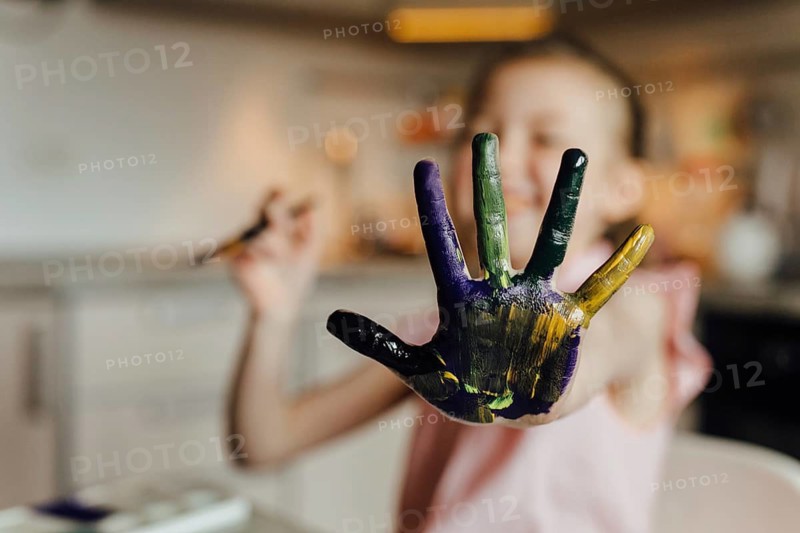 A joyful child showcases a hand covered in vibrant paint, expressing creativity and fun in an indoor art setting. The blurred background adds focus to the colorful hand