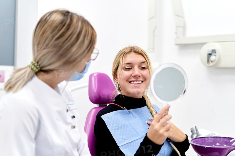 A confident woman smiles while holding a mirror during a dental check-up. A friendly dental professional in a mask discusses oral health in a modern, clean clinic setting