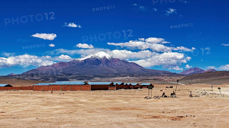 Lonely desert landscape with snow-covered mountain and blue sky in the background, lined with buildings, the town of Ollagüe in the Chilean Atacama Desert with the volcano of the same name Ollagüe in the background