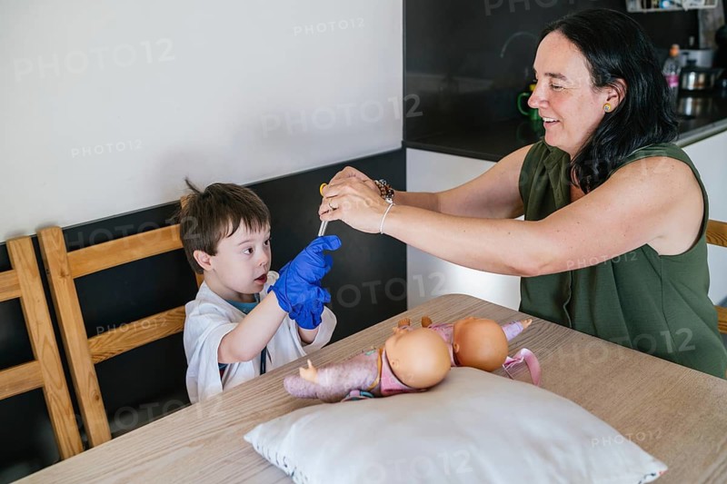 A mother and her son, who has Down syndrome, engage in imaginative play at the kitchen table. The boy, wearing a glove, focuses on the activity, demonstrating cooperation and creativity