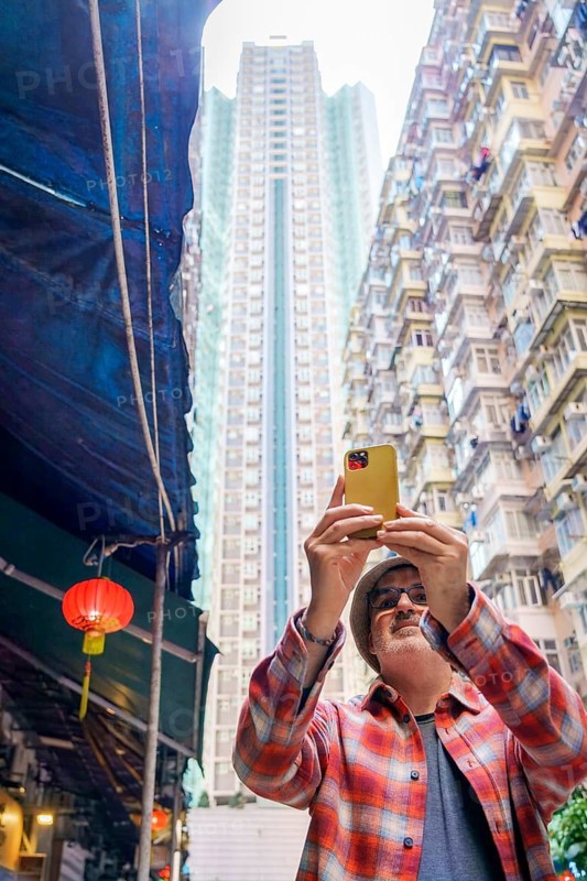 A middle-aged man uses his yellow smartphone to take photos of the bustling street scene in Hong Kong, showcasing a blend of traditional and modern elements with a red lantern and high-rise buildings