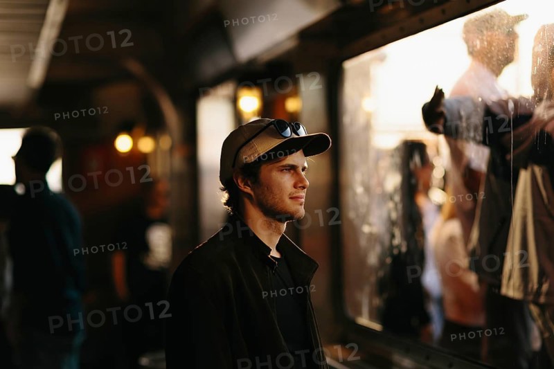 A young man in a casual cap gazes through a window, capturing a thoughtful moment amid the lively hustle of urban life in New York. The scene is warmly lit, enhancing the contemplative mood