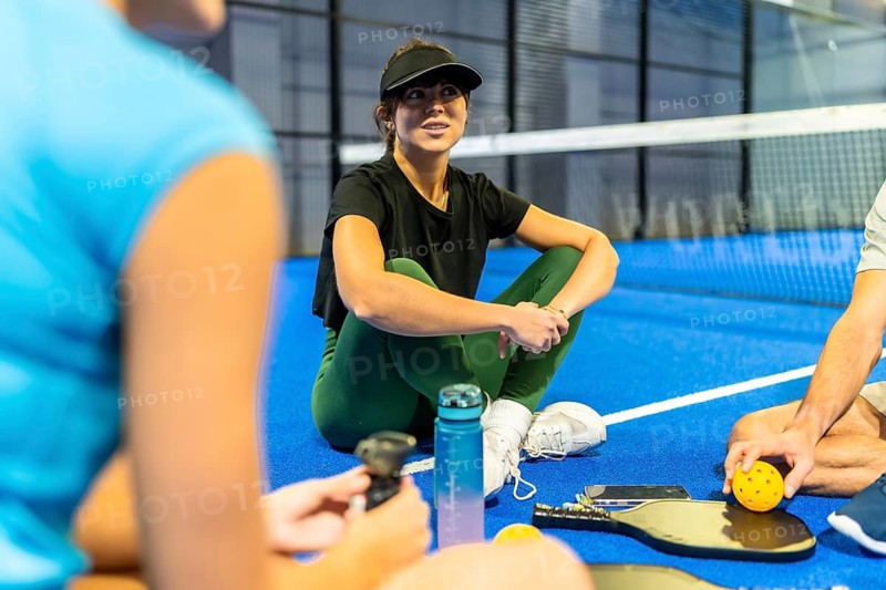 Young female athlete wearing a visor and athletic wear, sitting cross legged on a bright blue pickleball court, taking a break with teammates and holding equipment after playing