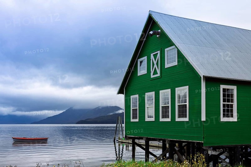 A vibrant green wooden house stands on stilts over a serene lake in Hoonah, Alaska, with misty mountains in the background and a small red boat floating nearby
