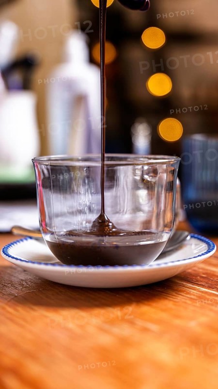 A stream of thick chocolate sauce flows into a clear glass cup set on a white saucer with a blue rim. Soft, warm cafe ambiance with blurred bokeh lights in the background