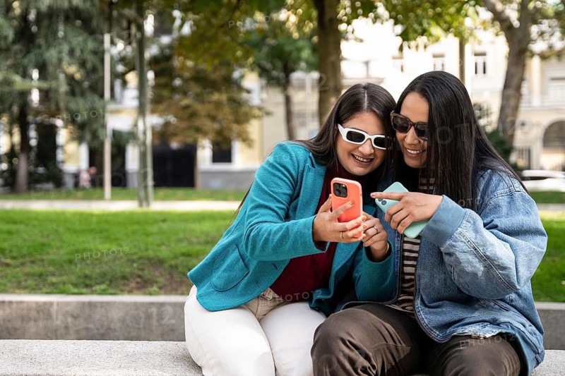 A joyful Latin lesbian couple sits on a park bench, sharing a smartphone. They wear casual outfits with sunglasses, embodying happiness and companionship in a green, urban setting