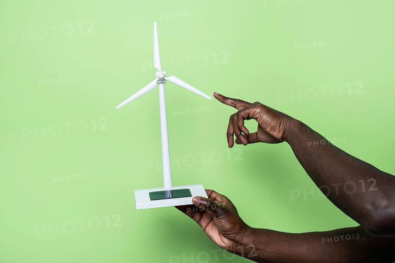 A hand delicately points at a miniature wind turbine on a green background, symbolizing sustainable energy and innovation. The setup emphasizes clean technology and environmental consciousness