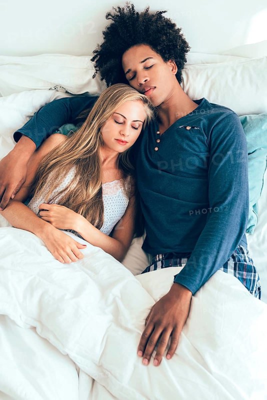 A serene couple is peacefully sleeping and embracing in a cozy bed. They radiate warmth and comfort, wrapped in a white blanket, depicting love and tranquility