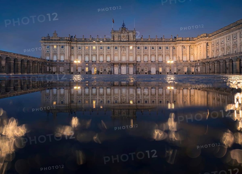 Stunning evening view of the Royal Palace of Madrid with illuminated windows, elegantly reflected in a water, creating a symmetrical and serene scene under a twilight sky