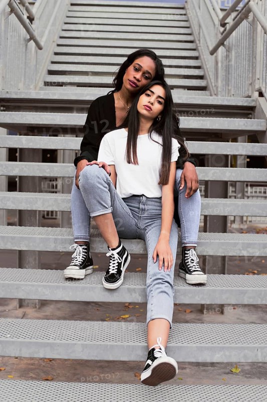 Two stylish friends relax on urban stairs, showcasing casual fashion. Jeans, t shirts, and sneakers highlight a laid back vibe in an outdoor city setting