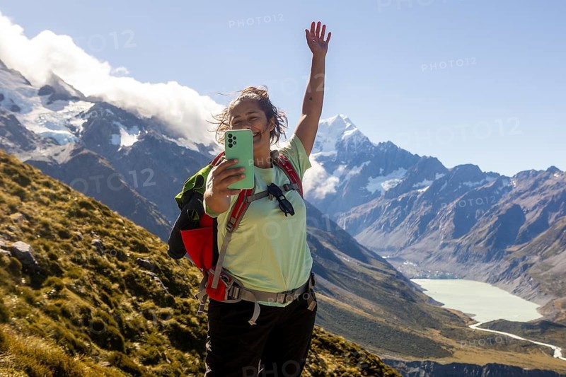 A woman is hiking on Mount Cook, New Zealand, during autumn. She joyfully takes a selfie with stunning mountains and a clear blue sky in the background, capturing the essence of adventure