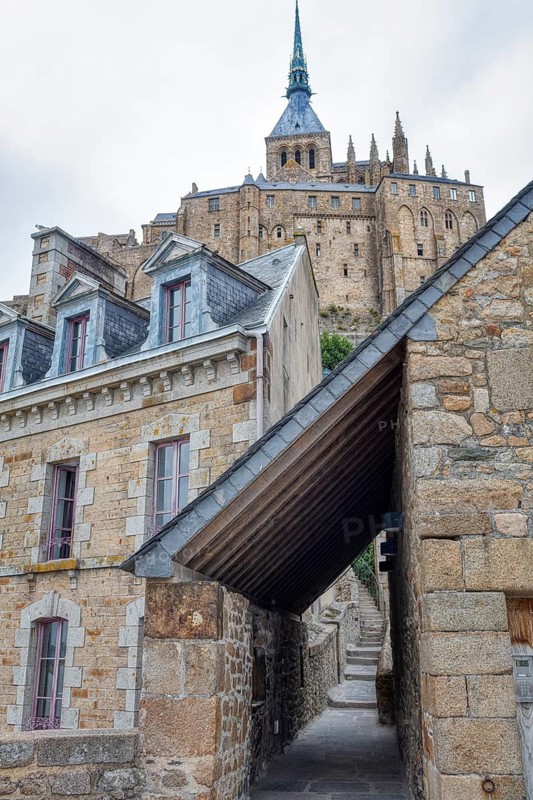 Historic stone buildings lining a narrow street with a church in the background, Le Mont-Saint-Michel