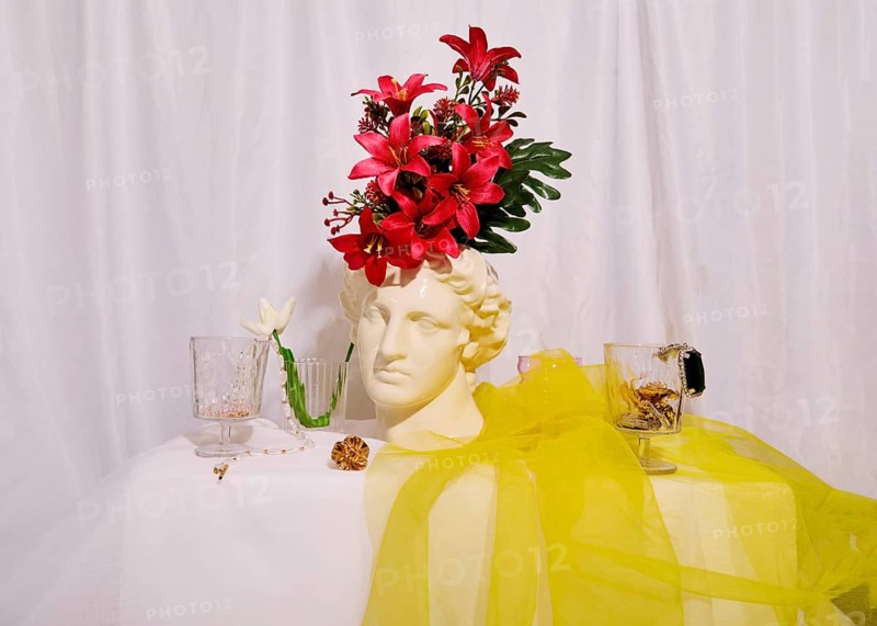 A contemporary still life scene featuring a classical bust adorned with vibrant pink flowers. Delicate glassware and a sheer yellow fabric drape the table, adding elegance