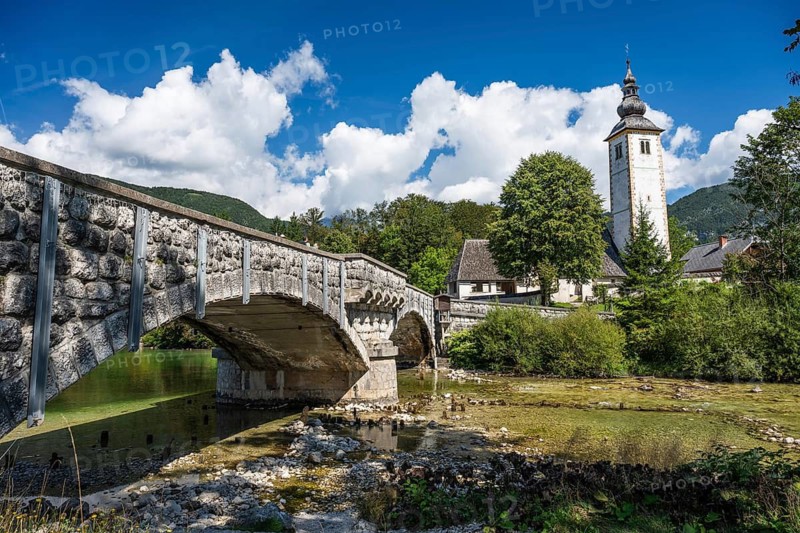 A picturesque view of the historic stone bridge and the Church of St. John the Baptist with its distinctive bell tower standing by the clear waters of Lake Bohinj in Slovenia