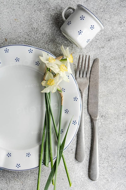 Top view of a charming table setting featuring a ceramic plate with blue dots, a matching mug, and fresh white daffodils laid beside stainless steel cutlery, all arranged on a textured gray background