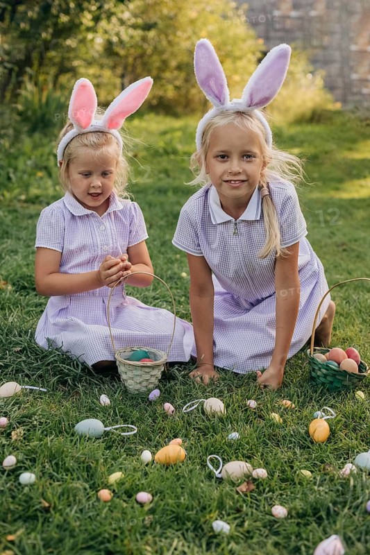 Two children wearing bunny ears participate in a joyful Easter egg hunt on a sunny day. They gather colorful eggs in baskets, surrounded by greenery and a cheerful atmosphere
