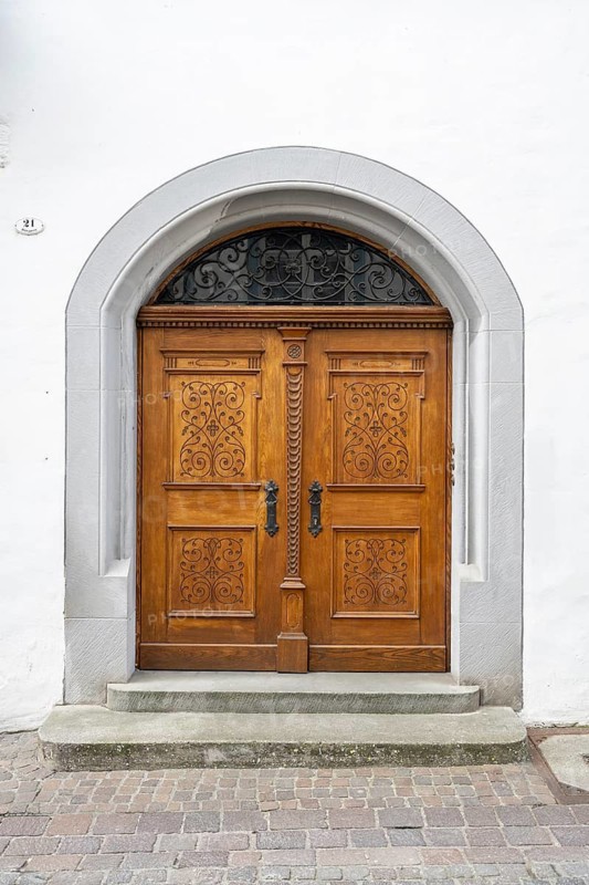 Frontal view of a historic portal on an old town house façade with a two-wing wooden door showing intricate carvings and floral ornaments in Baroque or Historicism styles, framed in a natural stone archway with a decorative skylight including wrought iron grille as an example of traditional craftsmanship. Konstanz, Konstanz District, Baden-Württemberg, Germany