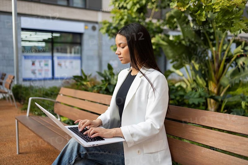 A professional woman in a white blazer works on a laptop, seated on a wooden bench in an urban park. Surrounded by greenery, she embodies a blend of business and lifestyle