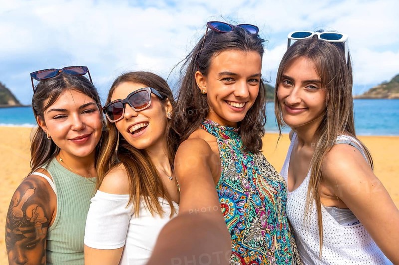 Group of happy young adult women enjoying vacation and friendship, posing for a selfie on a sunny beach with clear blue water and sky in the background