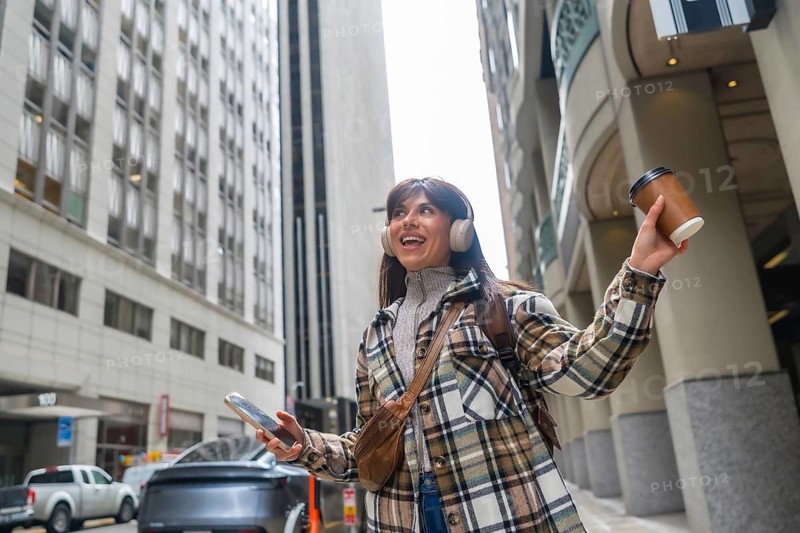 Young woman confidently walking along a city street, listening to music through headphones, holding takeaway coffee and a smartphone, enjoying her urban commute and active lifestyle
