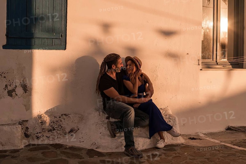 A couple embraces on a rustic Mediterranean street during sunset, casting long shadows on the whitewashed walls. The scene captures love and tranquility in Plaka, Milos island, Greece