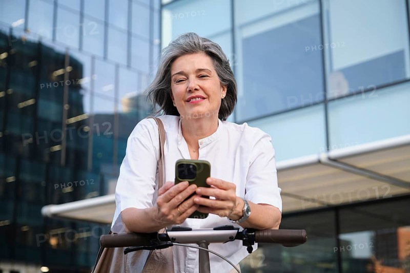 Mature woman in a stylish white blouse smiles while using a smartphone outdoors, leaning on an electric scooter, with a modern glass building in the background on a sunny day