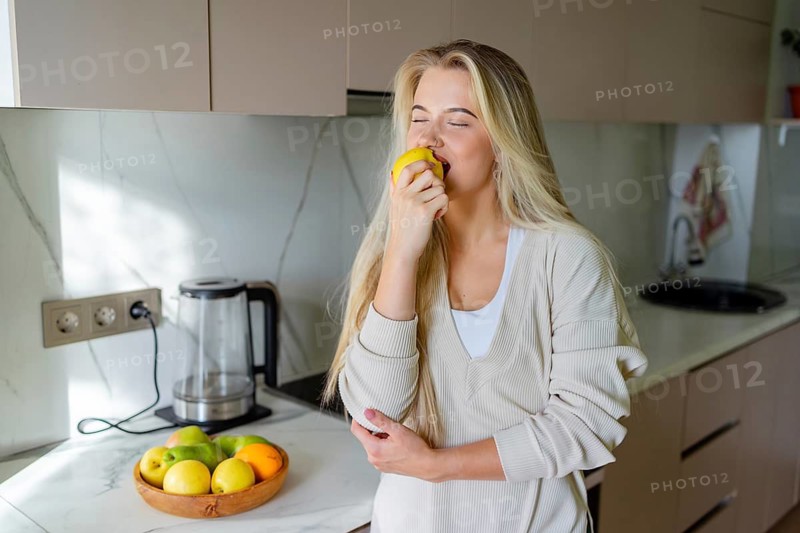 A woman stands in a stylish kitchen, savoring a juicy apple. Beside her is a bowl with various fruits, including apples, oranges and pears, enhancing the vibrant scene