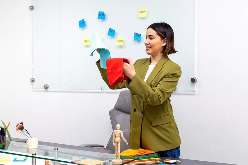 An image consultant stands in a modern office, holding up red and blue fabric swatches. The background shows a board with notes about fashion, style, and color coordination