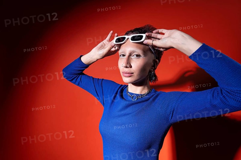 A stylish woman in a bright blue top poses confidently against a striking red background, adjusting her white sunglasses. The image captures a colorful and trendy fashion moment