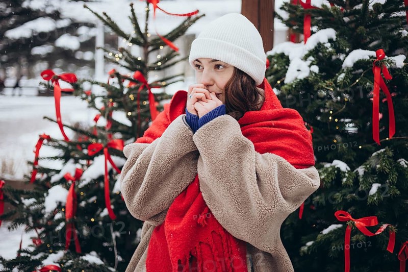 A young woman in a white beanie and red scarf stands cozily wrapped in a fluffy coat against a festive background with snow-covered Christmas trees decorated with red ribbons and lights