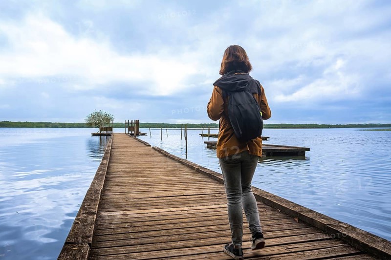 Woman backpacker walking on wooden pier into calm blue lake azur under cloudy sky, evoking travel, contemplation and peaceful escape in the natural landscape of landes, france