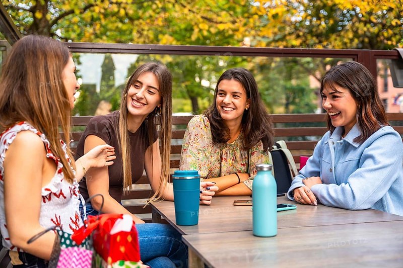 Group of smiling young female friends enjoying happy moments, communicating and laughing while sitting at a table outdoors in an urban cafe on an autumn day