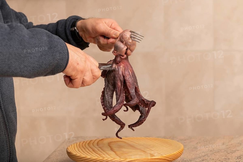 A man prepares a fresh octopus in the kitchen using a fork and scissors over a wooden cutting board. The background features neutral tiled walls and counter tops