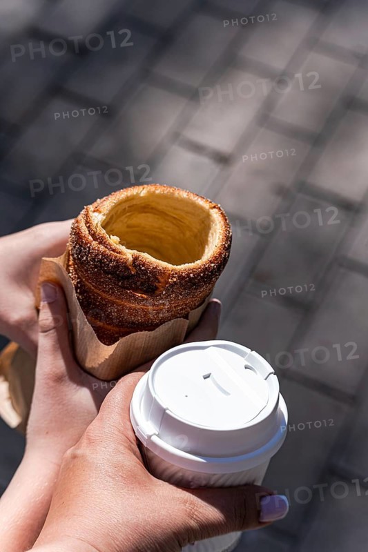 A boy holds a traditional trdelnik pastry in his hand, while a woman enjoys a cup of fresh coffee. The scene is set outdoors on a sunny day with a paved background