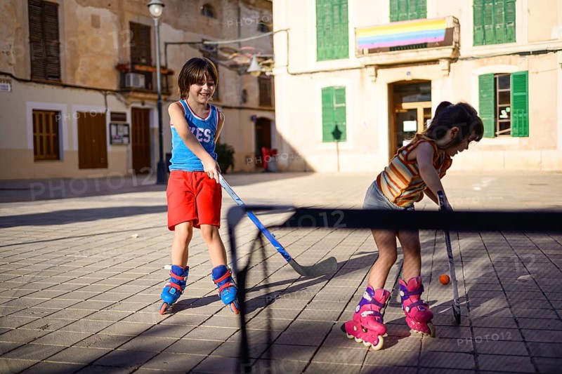 Two children enjoy street hockey on roller blades in a sunny plaza. Their vibrant outfits and high energy capture the joy and playfulness of outdoor activities