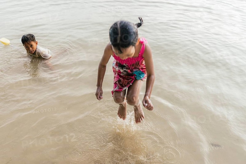 Children are playing joyfully at a beach in the Philippines. One child is jumping in shallow waters, while another is sitting, enjoying the calm sea