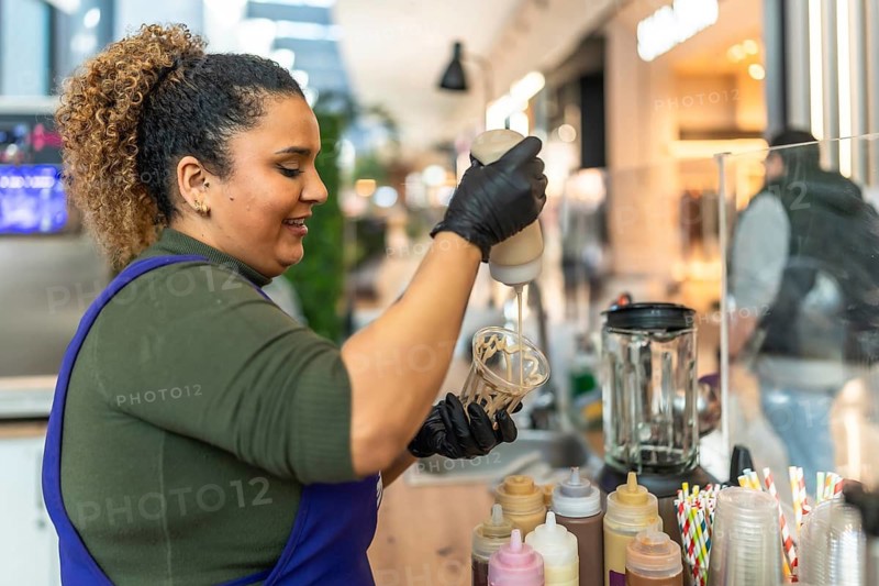 Smiling woman wearing black gloves and an apron, pouring syrup into a plastic cup for a milkshake, working at a fast food counter in a bustling commercial environment