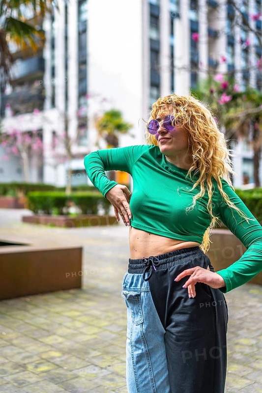 Caucasian woman with curly blonde hair and purple sunglasses dancing to modern music in a city park, wearing a green long sleeved top and two tone pants