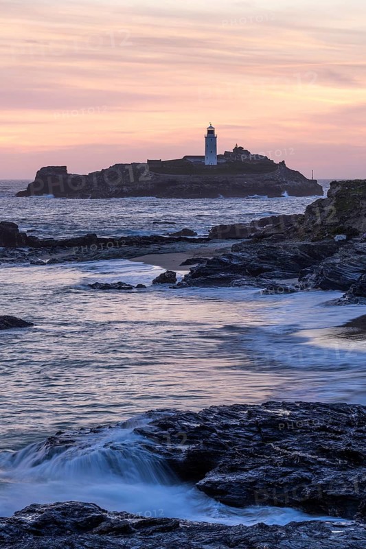 Godrevy Lighthouse, the lighthouse of Godrevy on Godrevy Island in the sea. Rocks in the foreground. In the evening at sunset with coloured sky. Cornwall, England, Great Britain