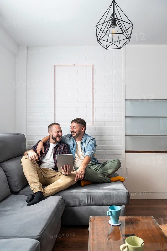 A joyful gay couple sits together on a comfortable couch in their modern living room, using a tablet and smiling at each other, creating a warm and inviting atmosphere