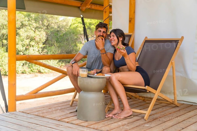 Young couple eating sandwiches and drinking coffee, sitting on wooden chairs on the terrace of their glamping tent, enjoying a relaxing vacation surrounded by nature