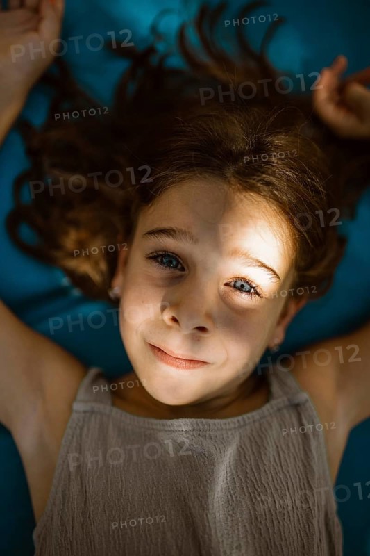 A child lies on a blue surface, smiling softly as sunlight casts delicate shadows on their face. Hair spread around creates a tranquil and joyful atmosphere