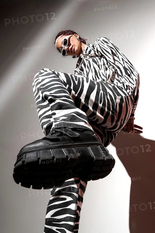 A fashion model in a striking zebra print outfit poses confidently in a studio. The upward angle highlights bold footwear and sunglasses, captured with flash photography