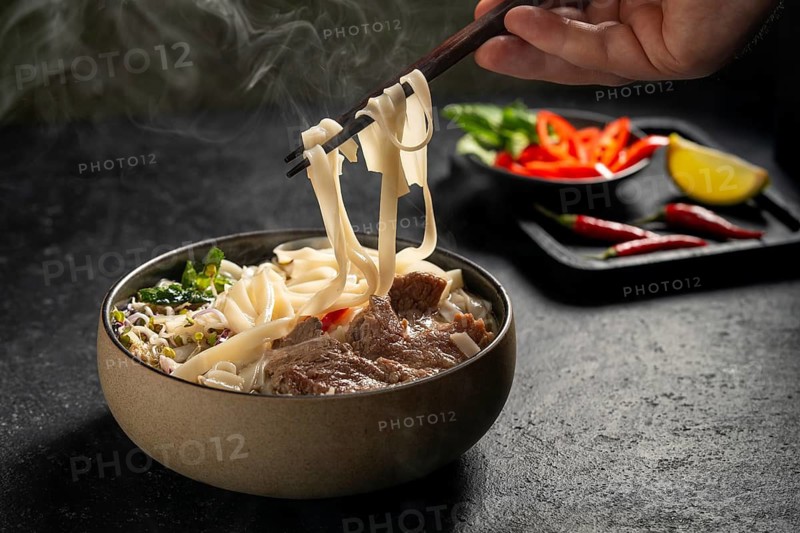 Cropped unrecognizable person's hand is seen using chopsticks to lift steamy, flat rice noodles from a bowl of Vietnamese beef pho. This aromatic soup includes tender slices of beef, onions, and a variety of fresh herbs, providing a savory experience