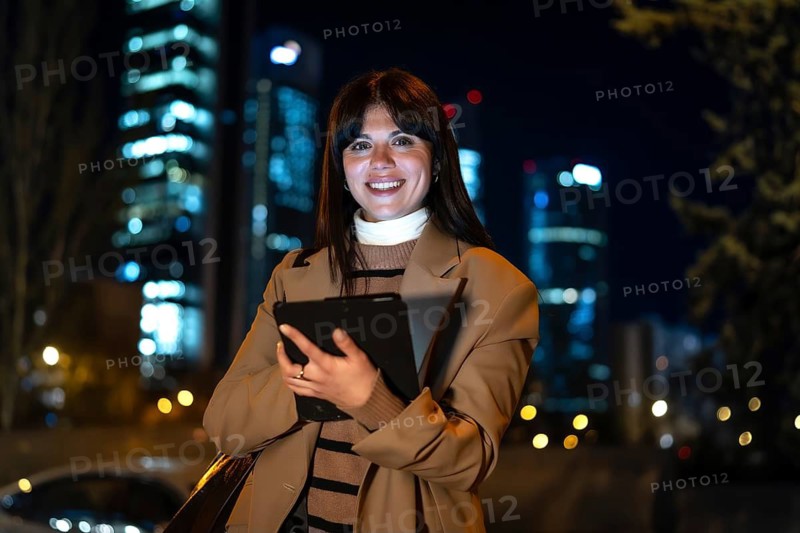 Young professional businesswoman smiling, holding a digital tablet outdoors at night in a city featuring illuminated skyscrapers, representing modern urban lifestyle and connectivity