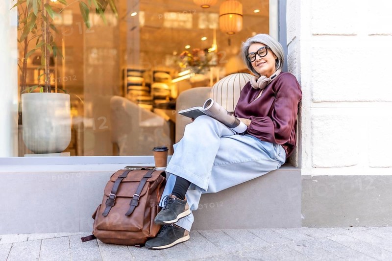 Active mature woman with gray hair wearing glasses and headphones, sitting on a window ledge, holding a magazine while relaxing outdoors with a takeaway coffee and backpack